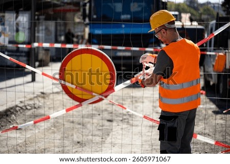 A construction worker in a hard hat and reflective vest is securing a safety barrier at a roadwork site. The setting is busy and well-lit, indicating a diligent effort to ensure safety.