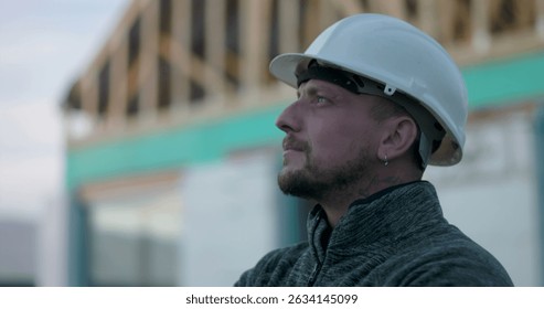 Construction worker in hard hat, side profile view, looking upward with focus at construction site, background of open frame structure, representing commitment and vision