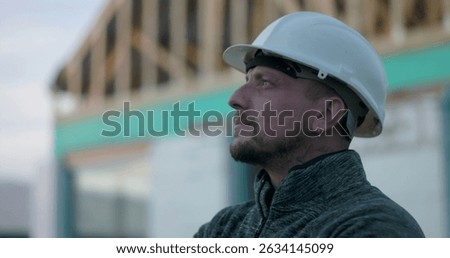 Construction worker in hard hat, side profile view, looking upward with focus at construction site, background of open frame structure, representing commitment and vision