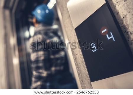A construction worker in a hard hat stands by an elevator display indicating floors three and four during a busy workday.