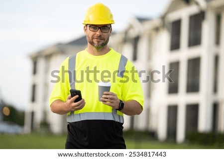 Construction worker having coffee break on construction site. A man builder in a helmet and goggles drinks a hot drink. Mature construction worker taking a break on site and drinking a coffee beverage