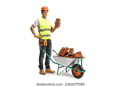 Construction worker holding a brick and standing behind a wheelbarrow with pile of bricks isolated on white background