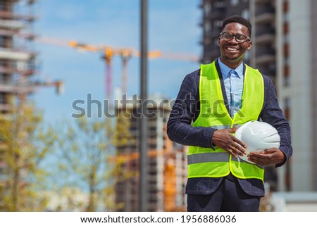 Construction Worker holding helmet at construction site. Worker with white safety helmet and yellow vest. Construction and industrial site workers concept 