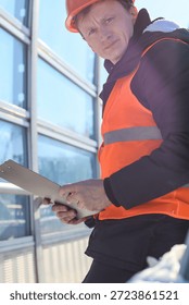 Construction worker inspecting clipboard documents. Worker wearing hard hat reviews paperwork on clipboard outside modern glass structure