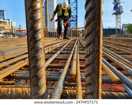 Construction worker inspecting steel reinforcement bars at a construction site. Close-up view of rebar with cranes and buildings in the background. Construction and engineering concept.