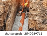 Construction worker installing drainage pipe in a trench during daylight, surrounded by gravel and soil