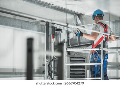 A construction worker is installing equipment while wearing a hard hat and safety harness in an industrial location, ensuring proper safety measures are in place.