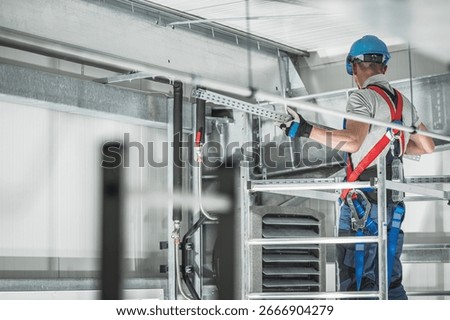 A construction worker is installing equipment while wearing a hard hat and safety harness in an industrial location, ensuring proper safety measures are in place.