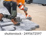 A construction worker is kneeling on the ground, carefully placing paving stones in a driveway. Wearing gloves and sturdy boots, they focus on arranging the stones