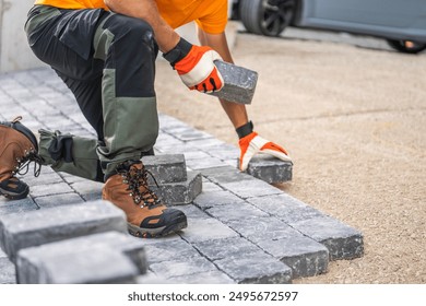 A construction worker is kneeling on the ground, carefully placing paving stones in a driveway. Wearing gloves and sturdy boots, they focus on arranging the stones - Powered by Shutterstock