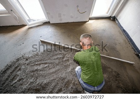 Construction worker leveling fresh semi-dry floor screed mixture of cement and sand over distribution heat system for underfloor heating in residential building is under construction.