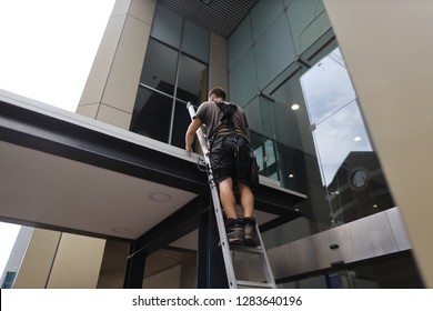 Construction Worker Maintenance Wearing Safety Harness Standing On The Industrial Ladder Repairing Roof Leaking, Sydney CBD, Australia   