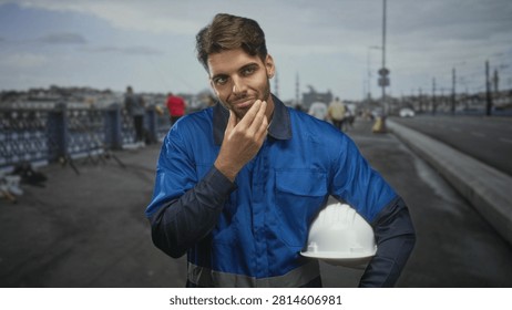 Construction worker man with beard touching chin while holding white hardhat and wearing blue overalls standing on street bridge by railing; contemplation pride.