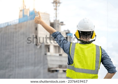 Construction worker man with green reflective vest and safety helmet showing thumbs up at unfinished building structure and tower crane. Male site engineer showing hand gesture sign for approval