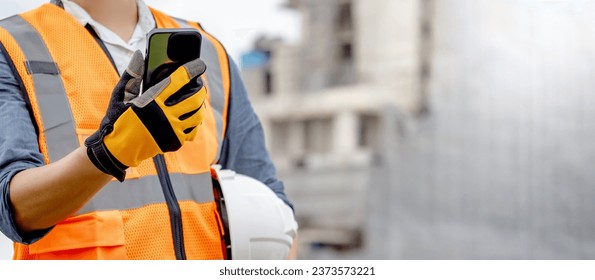 Construction worker man with orange reflective vest holding white protective safety helmet using smartphone at unfinished building site. Male engineer or foreman stay connected with social media app - Powered by Shutterstock