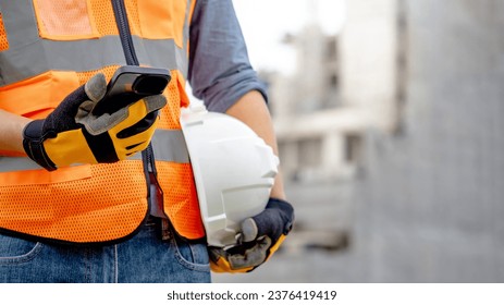 Construction worker man with orange reflective vest holding white protective safety helmet using smartphone at unfinished building site. Male engineer or foreman stay connected with social media app - Powered by Shutterstock