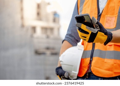 Construction worker man with orange reflective vest holding white protective safety helmet using smartphone at unfinished building site. Male engineer or foreman stay connected with social media app - Powered by Shutterstock