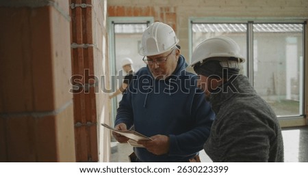 Construction worker measuring brick wall with engineer observing, teamwork in action at construction site with exposed walls and large windows