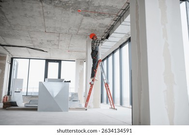 Construction worker on a ladder installing ventilation in a building under construction, wearing safety helmet and work overalls