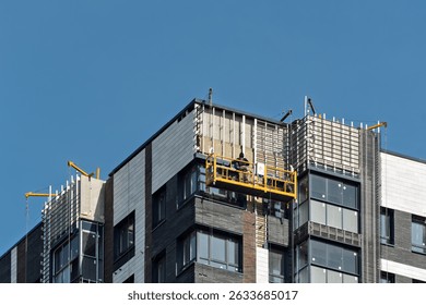 Construction worker on suspended platform repairs high-rise building facade against clear blue sky, modern urban development scene.