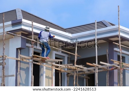 Construction Worker on wooden Scaffolding is Painting Roof structure of modern House against blue sky background
