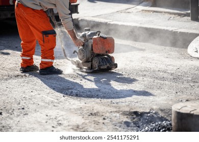 A construction worker operates a plate compactor on an outdoor site, creating dust as they prepare the surface for asphalt. The bright orange clothing stands out in the sunlight.