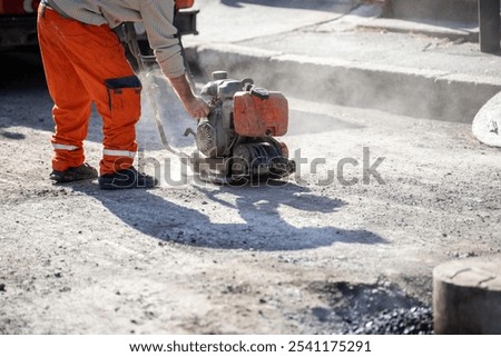 A construction worker operates a plate compactor on an outdoor site, creating dust as they prepare the surface for asphalt. The bright orange clothing stands out in the sunlight.