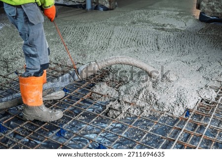 A construction worker in orange boots and gloves pours concrete over a rebar grid at a construction site. The worker is leveling the concrete to ensure an even surface.