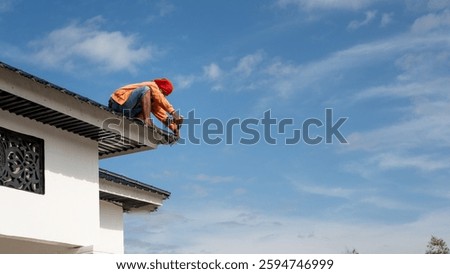 A construction worker in an orange jacket and red headscarf repairs a house roof using a power drill. The bright blue sky with scattered clouds contrasts with the worker's focused action.