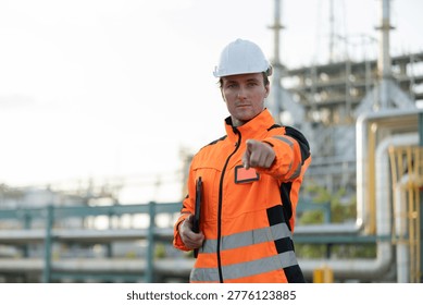 A construction worker in an orange safety jacket and white helmet points forward while holding a tablet at an industrial site.