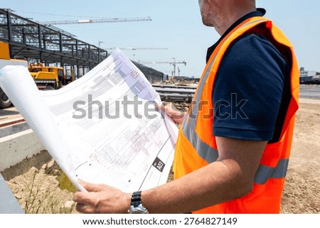 Construction worker in an orange vest examines blueprints at a construction site with cranes and steel structures in the background.