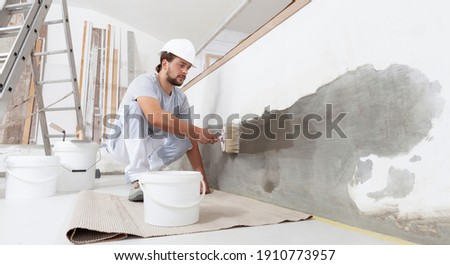 construction worker painter man with protective helmet, brush in hand and buckets of products to restore and paint the wall, indoor the building site of a house