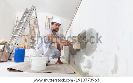 construction worker painter man with protective helmet, brush in hand and buckets of products to restore and paint the wall, indoor the building site of a house