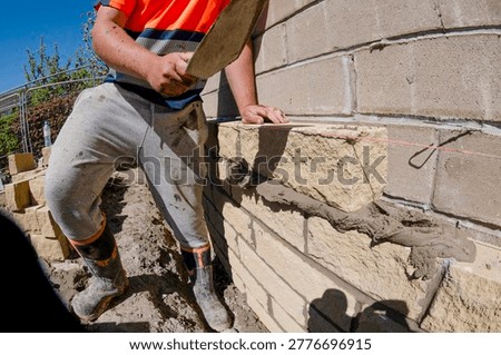 A construction worker is placing bricks on a wall at a job site. The worker uses a trowel and follows a guideline. The sun is shining in the background.
