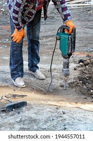 A Construction Worker In A Plaid Work Shirt And Blue Jeans Loosens Dense Soil With An Electric Jackhammer At A Construction Site, Copy Space, Vertical Image, Close-up.