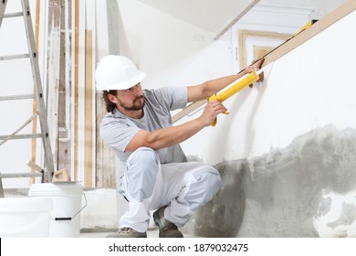 Construction Worker Plasterer Man Uses Caulking Gun In Building Site Of Home Renovation With Tools And Building Materials On The Floor