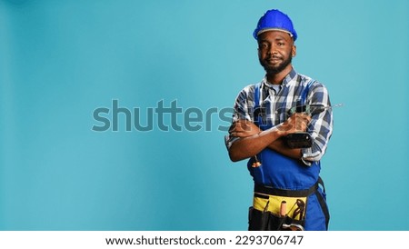 Construction worker posing with power drill gun, wearing hadhat and overalls in studio. Male builder holding electric nail drilling instrument, cordless machine equipment over background.