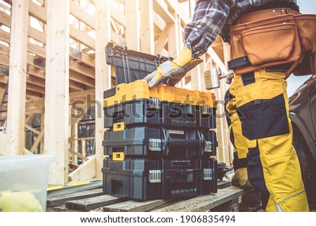 Construction Worker Preparing His Large Plastic Heavy Duty Toolboxes. Wooden Skeleton Frame of House in a Background. Construction Zone Theme.