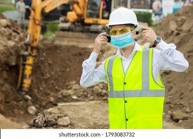 A Construction Worker Puts On A Face Mask To Protect Himself From Covid-19 With An Excavator In The Background