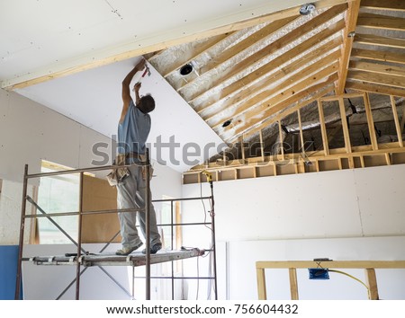 Construction Worker is putting up sheet rock. Hammering a nail into the sheetrock while on his tip toes.