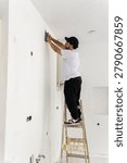 Construction worker reaching high on interior wall with finishing tool while standing on ladder in bright newly renovated room