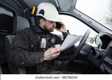 Construction worker reading papers, driving a car and talking on the phone while drinking coffee in Finland. He is wearing a white helmet and he has a dirty overalls.