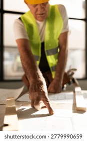 A construction worker in a reflective vest studies blueprints laid on a table. He points at specific sections while focused on the plans in a bright workspace.