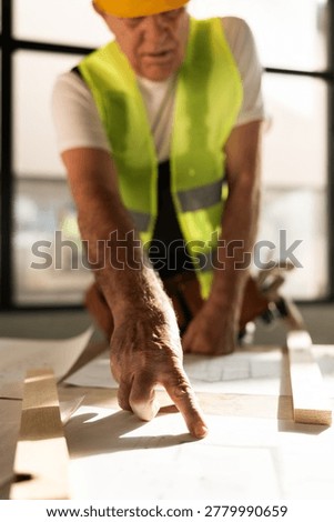 A construction worker in a reflective vest studies blueprints laid on a table. He points at specific sections while focused on the plans in a bright workspace.