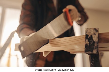 Construction worker renovating a home's interior kitchen. Using a crosscut saw, he cuts a wooden plank for molding installation. Contractor using his carpentry skills in a renovation project.