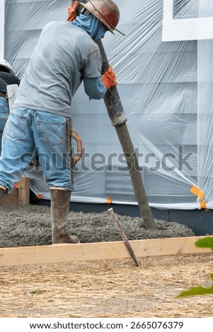 Construction worker in rubber boots and hard hat working cement pumper hose to pour fresh cement into wood forms for concrete back patio and stairs, new housing development

