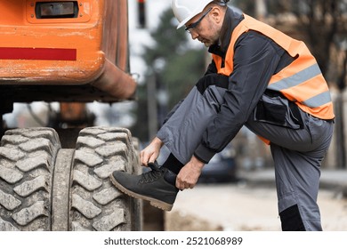 A construction worker in safety gear is kneeling to tie his boot next to large machinery on a bustling city street, preparing for the day's tasks. - Powered by Shutterstock