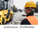 A construction worker in safety gear observes large machinery working on a city street, ensuring safety and efficiency in the project during daylight.