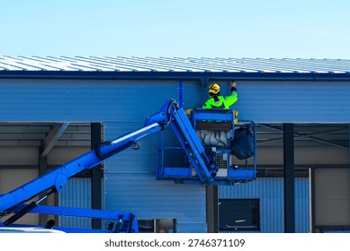 Construction worker in safety gear using boom lift to inspect and maintain metal roof of industrial warehouse