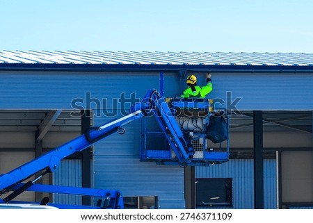 Construction worker in safety gear using boom lift to inspect and maintain metal roof of industrial warehouse